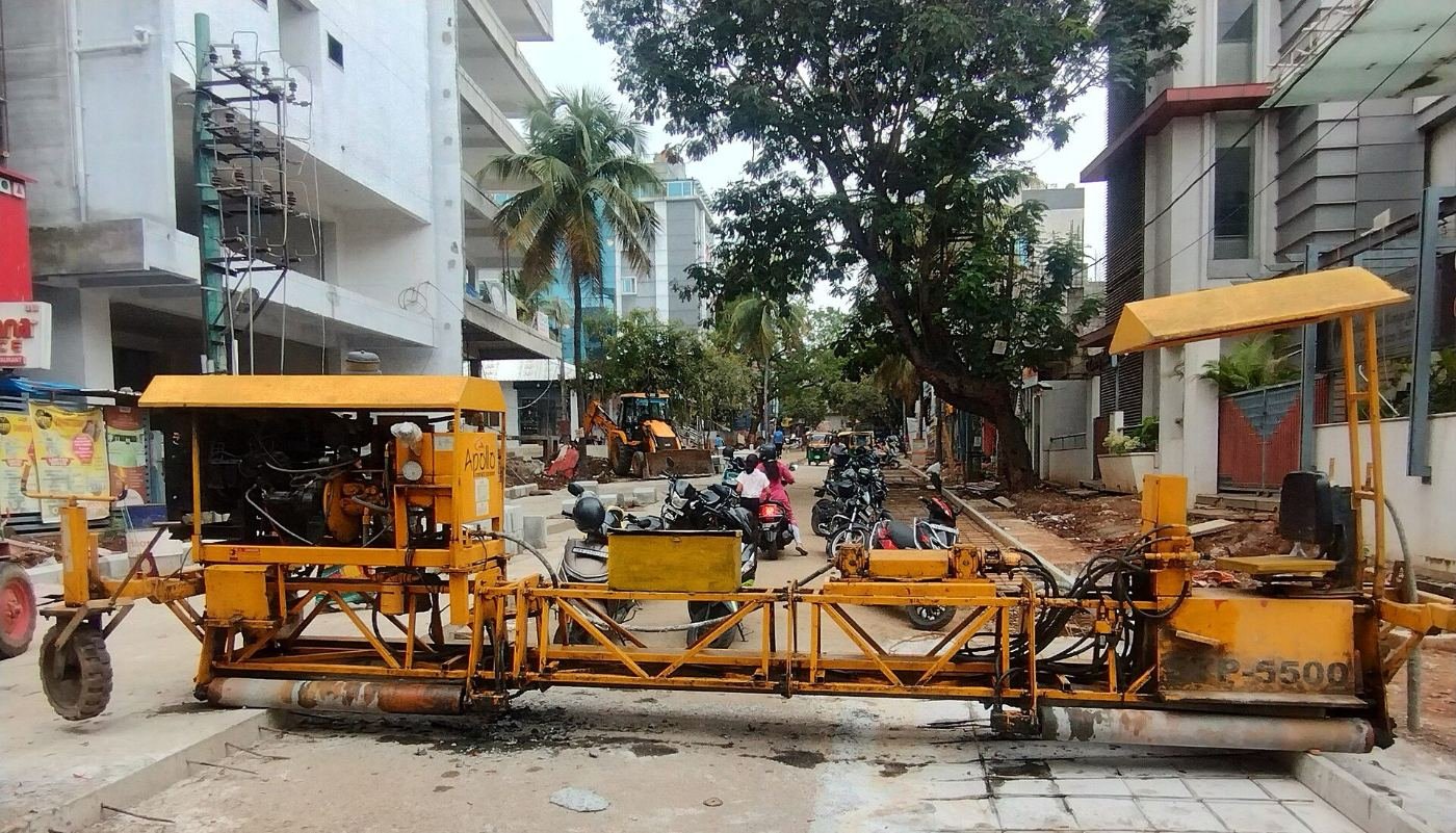 A yellow paver machine laying a road in RR Nagar, Bengaluru, with two-wheelers passing beside it